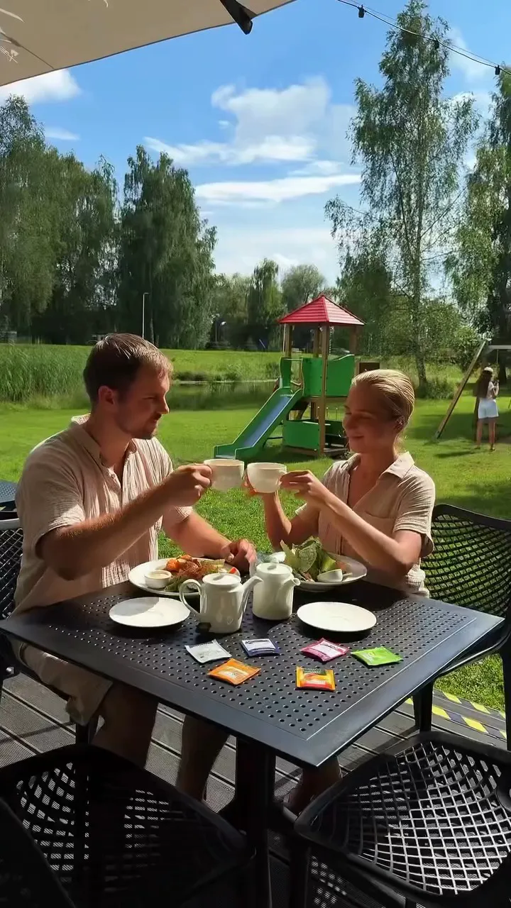 Divi viesi pie galdiņa terasē saskandina tējas tasītes, fonā zāliens un bērnu rotaļu laukums